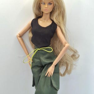 May include: A fashion doll with long blonde hair wearing a black sleeveless top and olive green cargo pants. The pants feature large side pockets and a thin, bright neon yellow drawstring cord tied at the waist. The doll has articulated limbs.