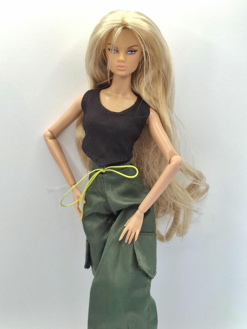 May include: A fashion doll with long blonde hair wearing a black sleeveless top and olive green cargo pants. The pants feature large side pockets and a thin, bright neon yellow drawstring cord tied at the waist. The doll has articulated limbs.