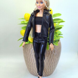 May include: A doll with blonde hair and blue eyes, wearing a black leather jacket, a black tube top, and black leather pants. The doll is also wearing black shoes. The doll is standing in front of a decorative pot with yellow fruit and green leaves.