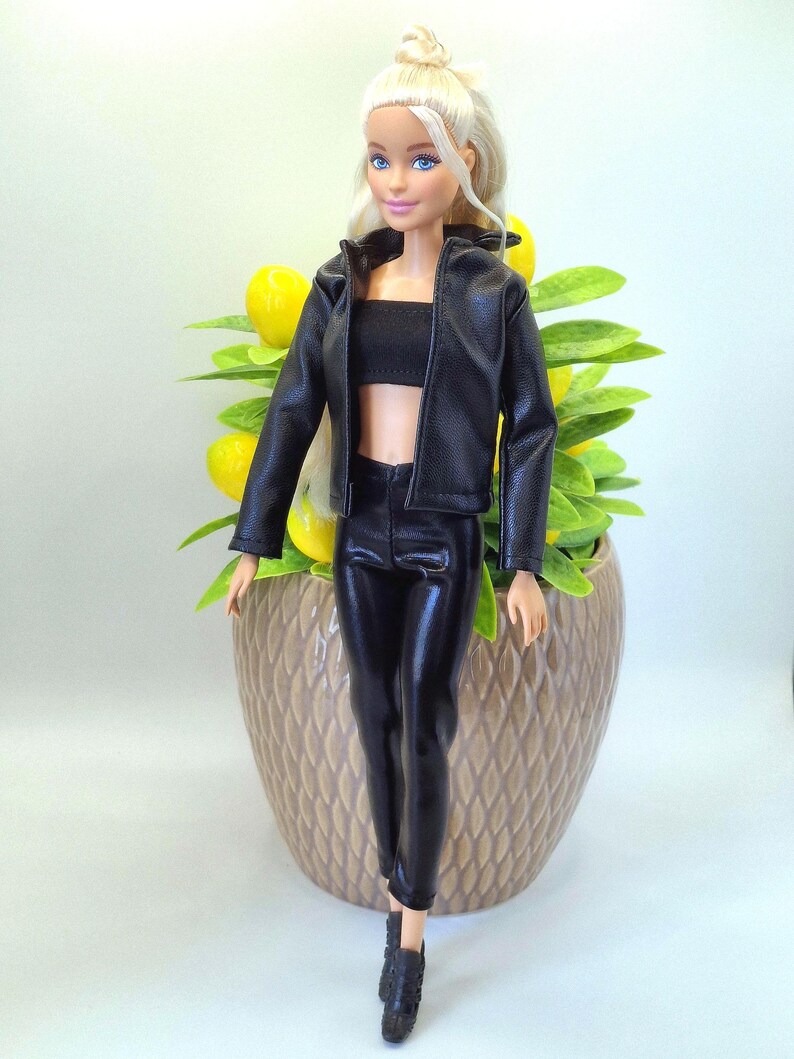 May include: A doll with blonde hair and blue eyes, wearing a black leather jacket, a black tube top, and black leather pants. The doll is also wearing black shoes. The doll is standing in front of a decorative pot with yellow fruit and green leaves.