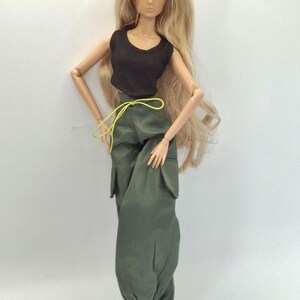 May include: A fashion doll with long blonde hair wearing a black sleeveless crop top and olive green cargo pants featuring side pockets, a neon yellow drawstring waist, and cinched ankles, paired with black boots.