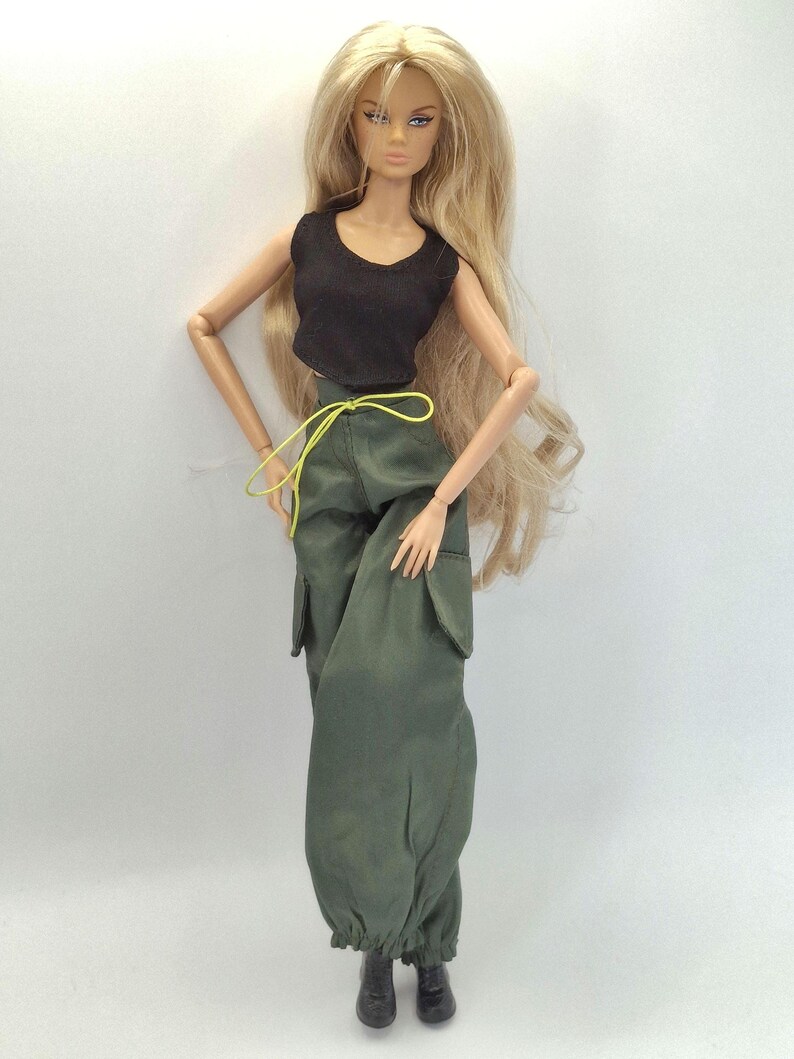 May include: A fashion doll with long blonde hair wearing a black sleeveless crop top and olive green cargo pants featuring side pockets, a neon yellow drawstring waist, and cinched ankles, paired with black boots.