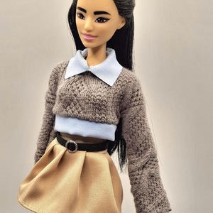 May include: A doll with long black hair wearing a gray knit sweater with a light blue collar and a tan pleated skirt. A black belt with a silver buckle cinches the waist. The doll has a neutral expression.