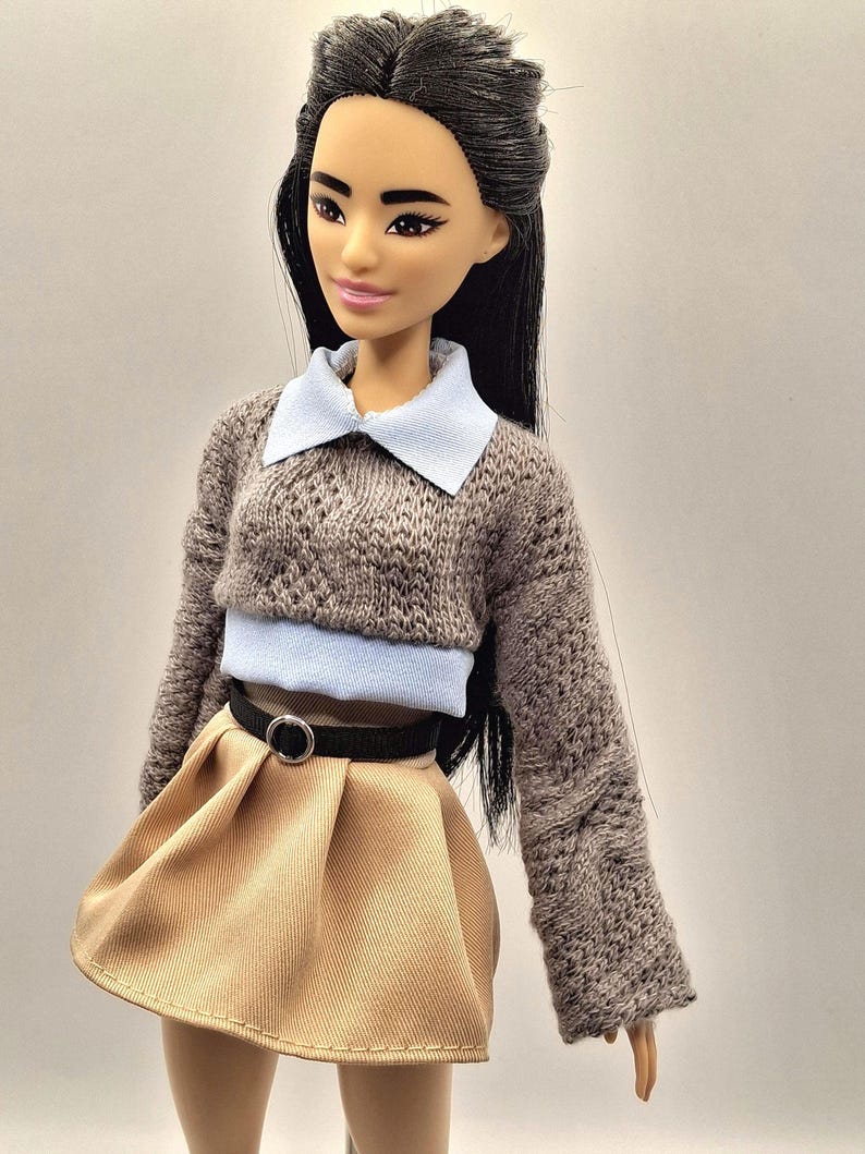 May include: A doll with long black hair wearing a gray knit sweater with a light blue collar and a tan pleated skirt. A black belt with a silver buckle cinches the waist. The doll has a neutral expression.