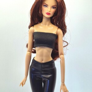 May include: A fashion doll with long auburn hair, wearing a black tube top and black pants. The doll has a fair complexion, red lipstick, and is posed against a plain background. The doll's outfit appears to be made of a shiny material.