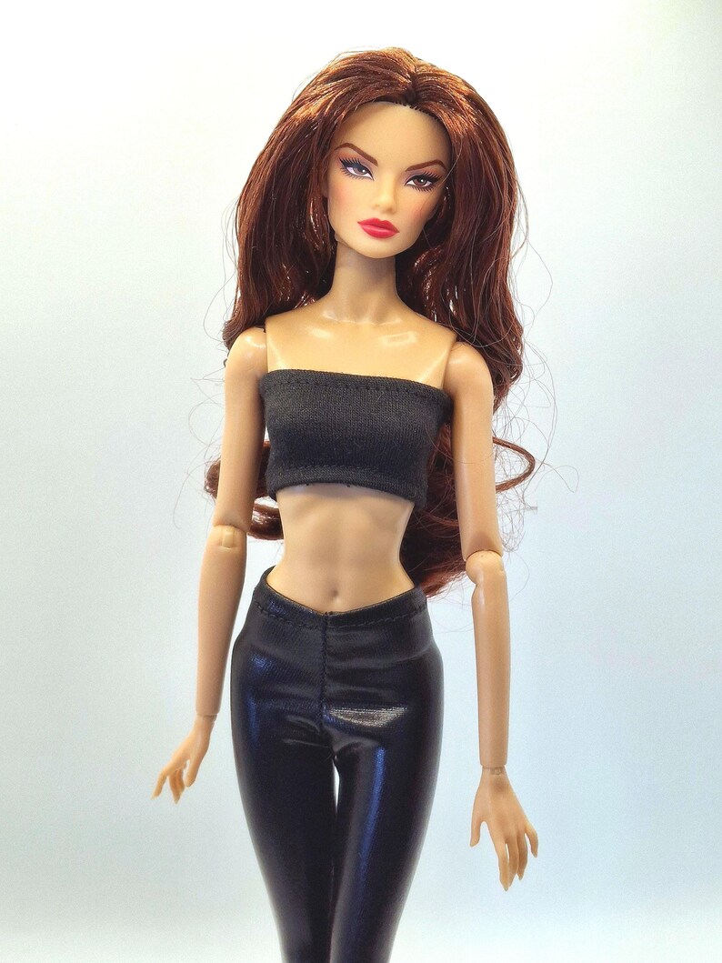 May include: A fashion doll with long auburn hair, wearing a black tube top and black pants. The doll has a fair complexion, red lipstick, and is posed against a plain background. The doll's outfit appears to be made of a shiny material.