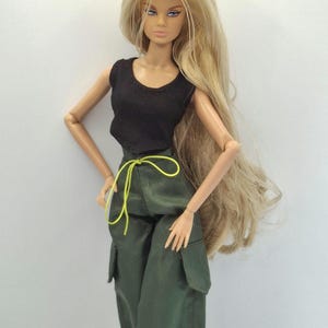 May include: A fashion doll with long blonde hair, wearing a black sleeveless top and olive green cargo pants featuring large side pockets and a bright neon yellow drawstring waist tie. The doll has articulated arms and detailed facial features.