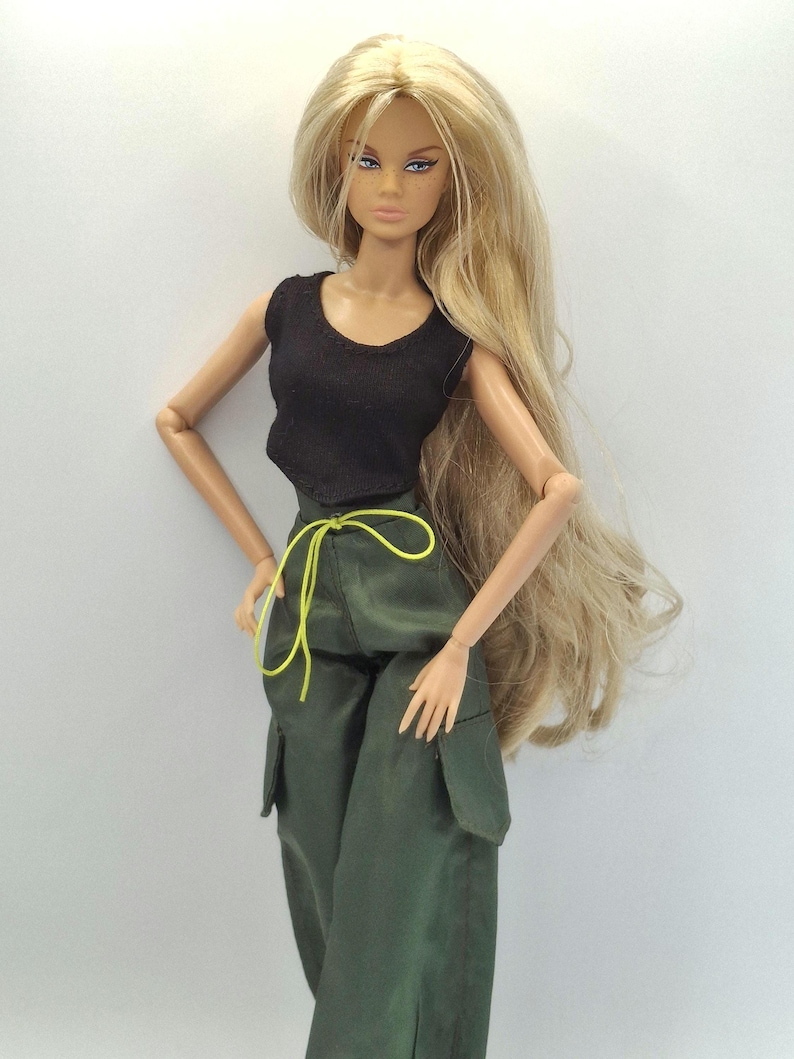 May include: A fashion doll with long blonde hair, wearing a black sleeveless top and olive green cargo pants featuring large side pockets and a bright neon yellow drawstring waist tie. The doll has articulated arms and detailed facial features.