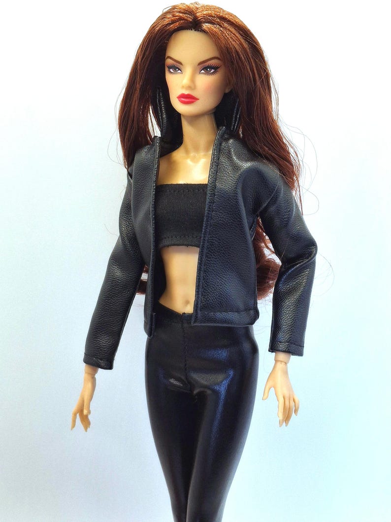 May include: A fashion doll with long auburn hair, wearing a black faux leather jacket, a black tube top, and black pants. The doll has red lipstick and is posed against a white background. The jacket is open, revealing the top and midriff.
