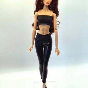 May include: A fashion doll with long auburn hair, wearing a black tube top and black leggings. The doll is posed with arms at its sides and is wearing black ankle boots. The doll has a fair complexion and is standing on a clear plastic base.