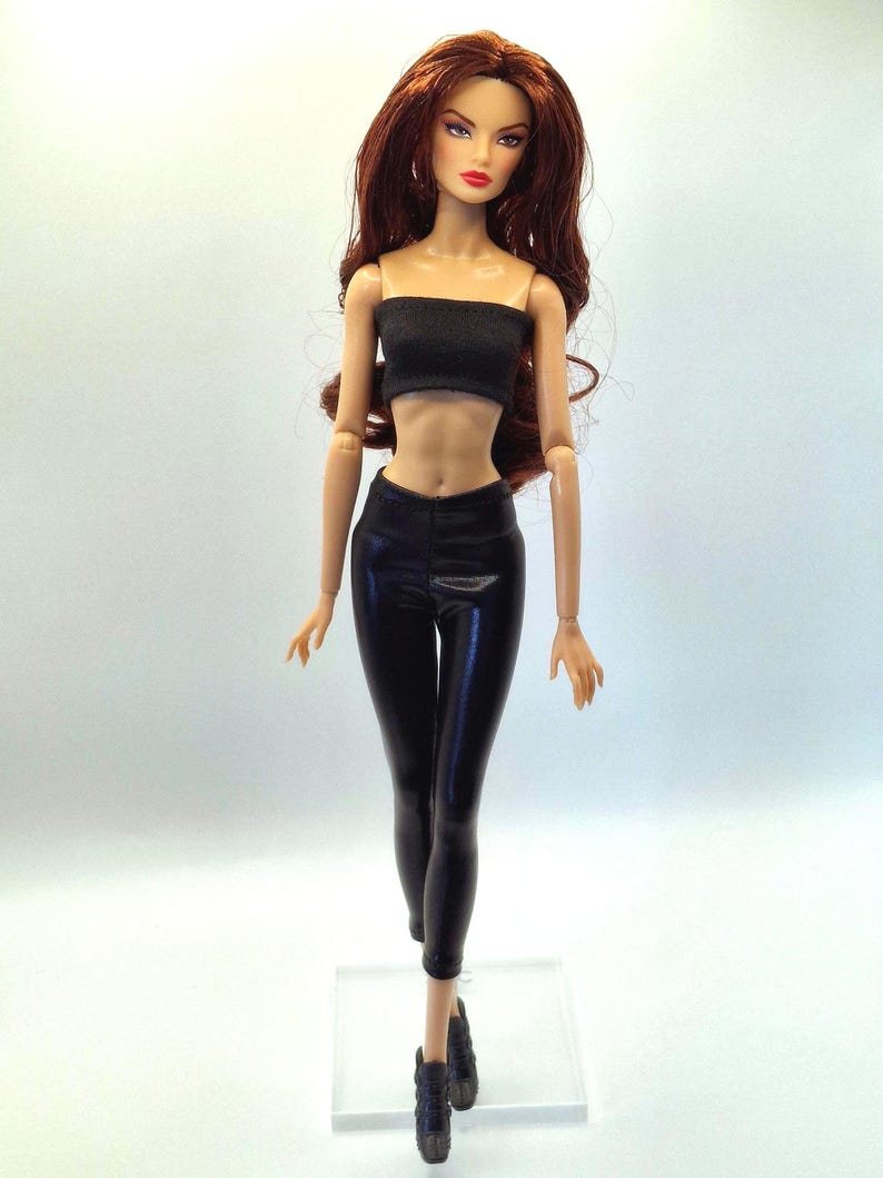 May include: A fashion doll with long auburn hair, wearing a black tube top and black leggings. The doll is posed with arms at its sides and is wearing black ankle boots. The doll has a fair complexion and is standing on a clear plastic base.