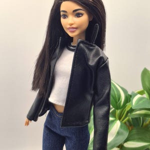 May include: A doll with long, dark hair wearing a black faux leather jacket over a white top and blue jeans. The doll has a neutral expression and is posed in front of a blurred green plant.