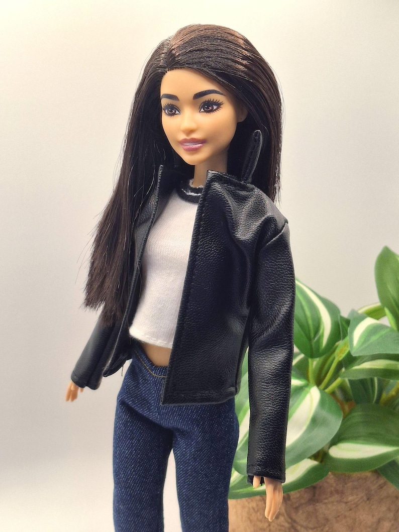 May include: A doll with long, dark hair wearing a black faux leather jacket over a white top and blue jeans. The doll has a neutral expression and is posed in front of a blurred green plant.
