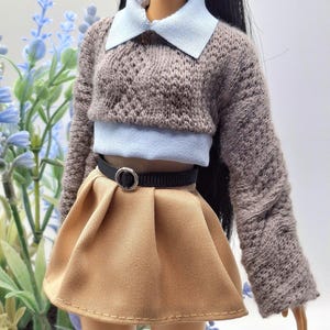 May include: A doll wearing a brown knit sweater with a light blue collar and a matching light blue top. The doll is also wearing a tan pleated skirt with a black belt. The outfit is a miniature fashion ensemble.