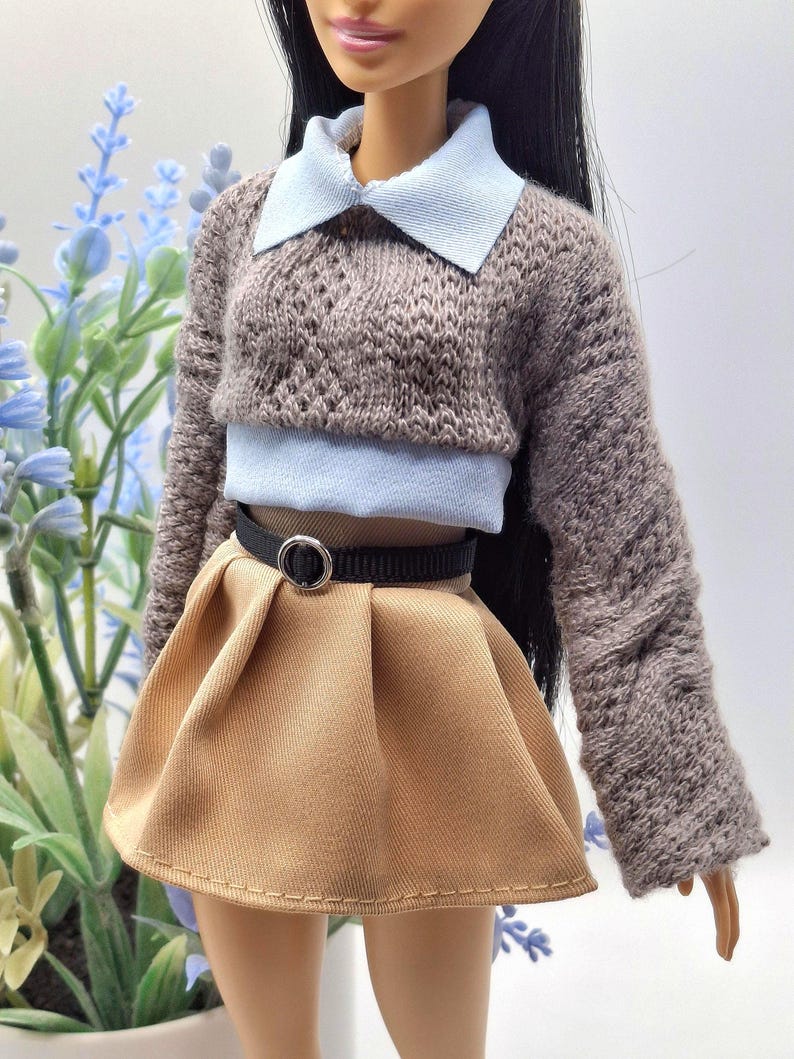 May include: A doll wearing a brown knit sweater with a light blue collar and a matching light blue top. The doll is also wearing a tan pleated skirt with a black belt. The outfit is a miniature fashion ensemble.