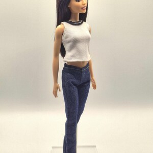 May include: A doll wearing a white sleeveless top with a black trim and dark blue denim jeans. The doll has long, dark hair and is wearing white sneakers. The doll is standing on a clear plastic stand.