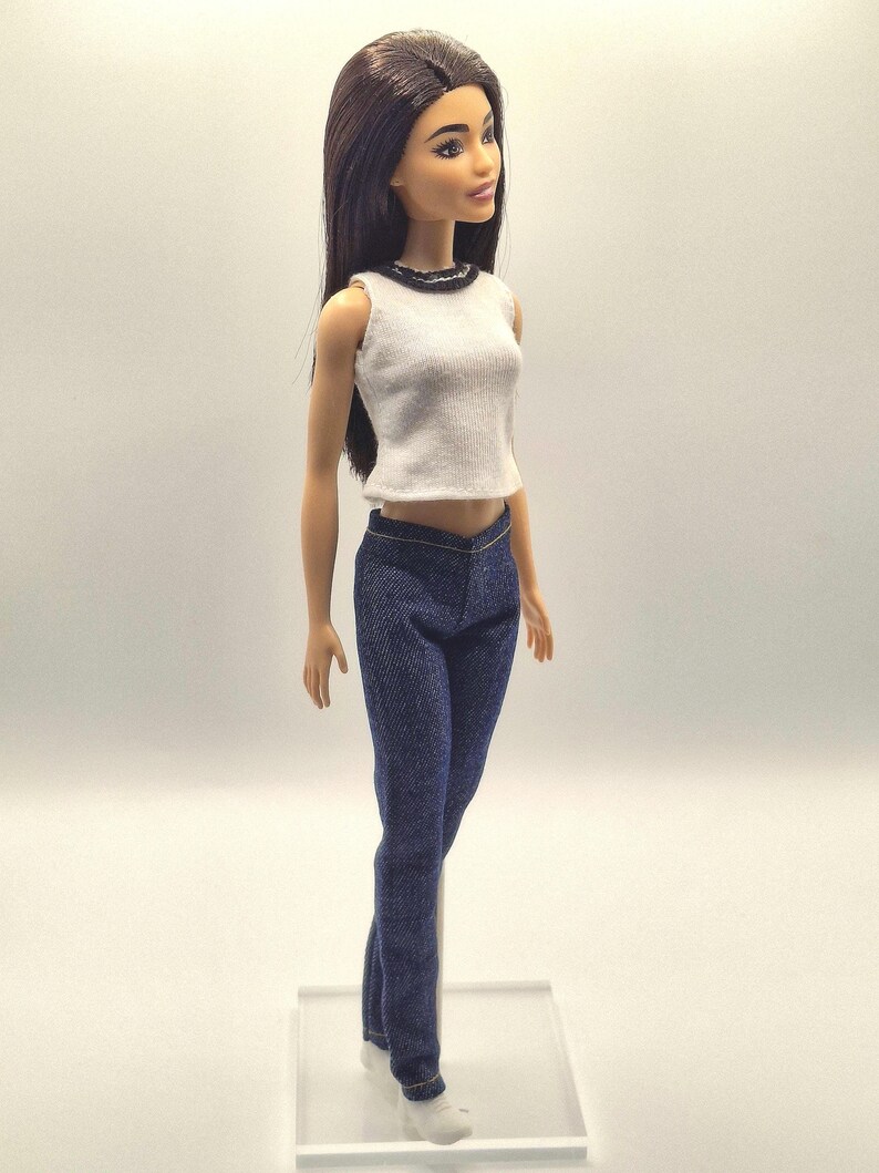 May include: A doll wearing a white sleeveless top with a black trim and dark blue denim jeans. The doll has long, dark hair and is wearing white sneakers. The doll is standing on a clear plastic stand.