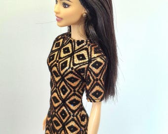 May include: A fashion doll wearing a black and gold geometric pattern mini dress with short sleeves. The doll has long, dark brown hair and a black headband. The doll is wearing small gold earrings.
