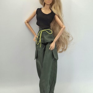 May include: A fashion doll with long blonde hair wearing a black sleeveless crop top and olive green cargo pants. The pants feature large side pockets, a bright yellow drawstring at the waist, and elasticated cuffs at the ankles.