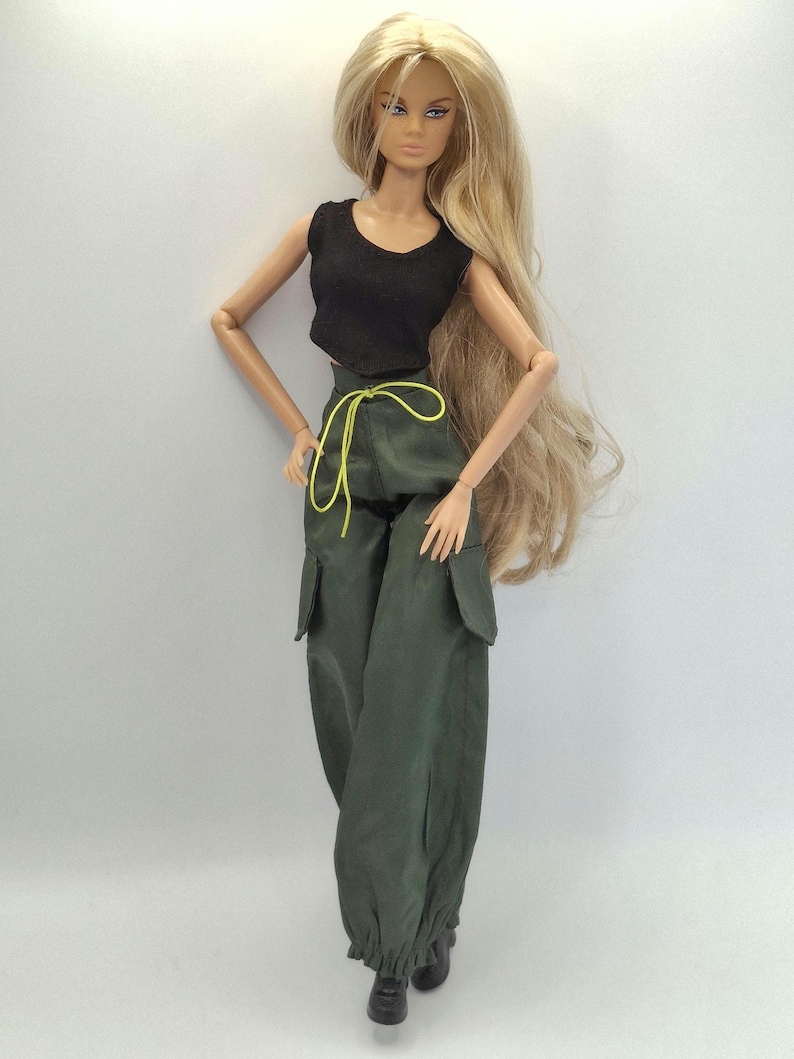 May include: A fashion doll with long blonde hair wearing a black sleeveless crop top and olive green cargo pants. The pants feature large side pockets, a bright yellow drawstring at the waist, and elasticated cuffs at the ankles.