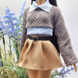 May include: A doll wearing a brown knit sweater with a light blue collared shirt underneath. The doll is also wearing a tan pleated skirt and a black belt with a silver buckle. The doll is posed in front of blue flowers.