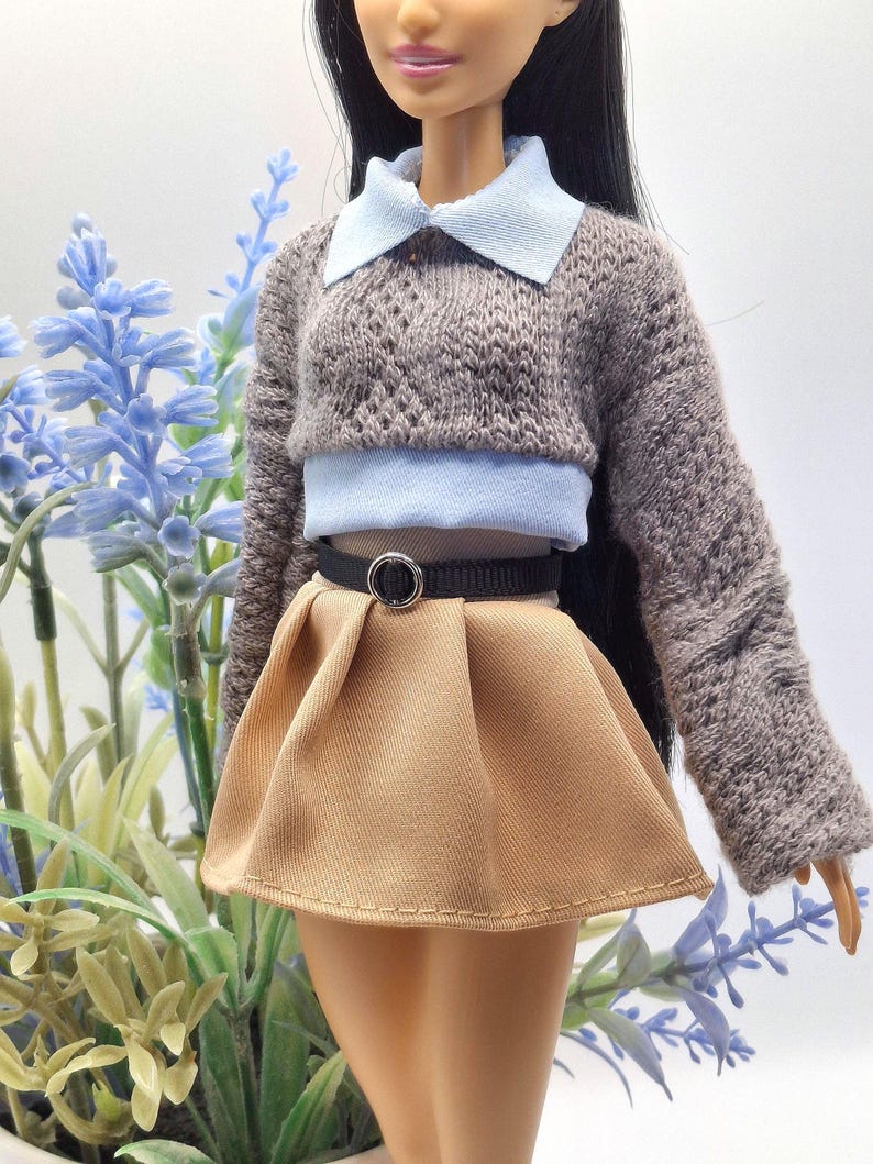 May include: A doll wearing a brown knit sweater with a light blue collared shirt underneath. The doll is also wearing a tan pleated skirt and a black belt with a silver buckle. The doll is posed in front of blue flowers.