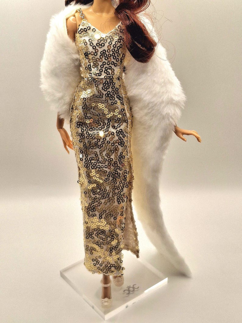 May include: A doll wearing a gold sequined gown with a slit, paired with a white faux fur wrap. The dress has thin straps and a V-neck. The doll has auburn hair and is standing on a clear stand.