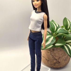 May include: A doll with long brown hair, wearing a white sleeveless top with a black trim and dark blue denim jeans. The doll is standing on a clear plastic stand, with white sneakers. A plant in a brown pot is in the background.