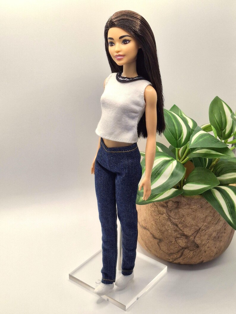 May include: A doll with long brown hair, wearing a white sleeveless top with a black trim and dark blue denim jeans. The doll is standing on a clear plastic stand, with white sneakers. A plant in a brown pot is in the background.