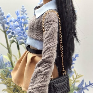 May include: A doll wearing a light blue collared shirt, a brown knit sweater, and a tan pleated skirt. A black belt and a black quilted purse with a gold chain complete the outfit. The doll has long black hair and a silver earring.