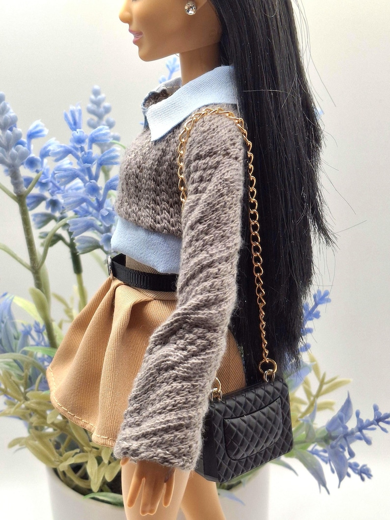 May include: A doll wearing a light blue collared shirt, a brown knit sweater, and a tan pleated skirt. A black belt and a black quilted purse with a gold chain complete the outfit. The doll has long black hair and a silver earring.