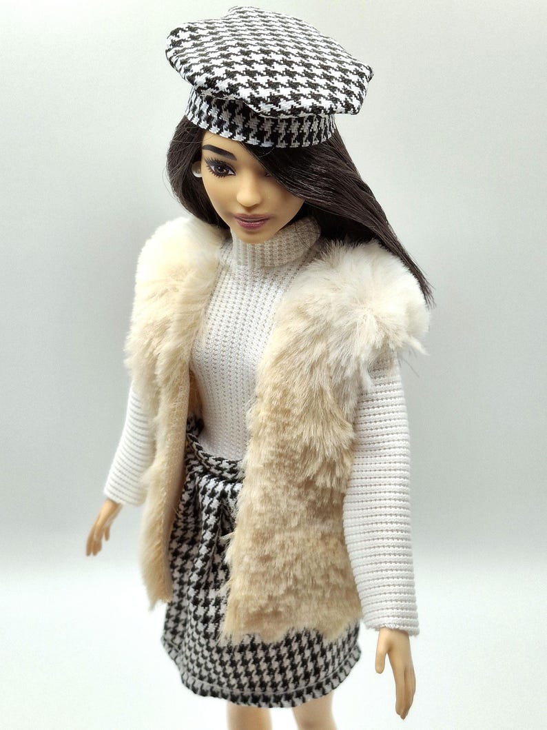 May include: A doll wearing a white turtleneck sweater, a beige faux fur vest, and a black and white houndstooth skirt. The doll also wears a matching houndstooth beret. The doll has dark hair and is posed against a white background.