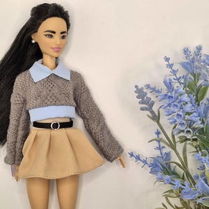 May include: A doll wearing a gray sweater with a light blue collar and a tan skirt. The doll has long black hair and is accessorized with a black belt. Blue flowers are in the background.