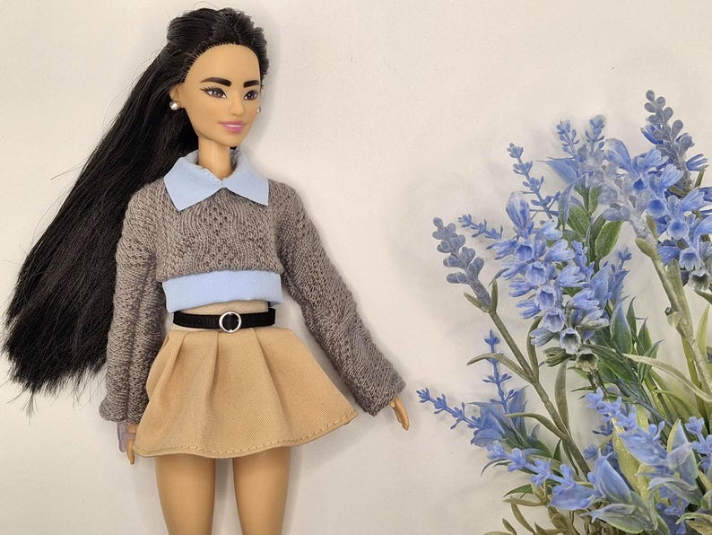 May include: A doll wearing a gray sweater with a light blue collar and a tan skirt. The doll has long black hair and is accessorized with a black belt. Blue flowers are in the background.