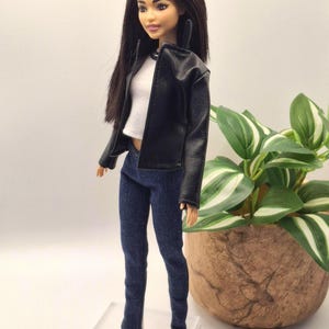 May include: A fashion doll wearing a black faux leather jacket, white top, and dark blue denim jeans. The doll has long dark hair and is wearing white shoes. The doll is standing on a clear plastic stand.