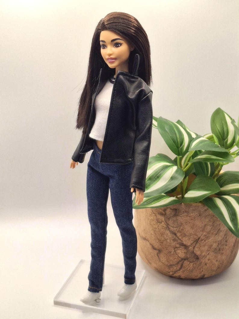 May include: A fashion doll wearing a black faux leather jacket, white top, and dark blue denim jeans. The doll has long dark hair and is wearing white shoes. The doll is standing on a clear plastic stand.
