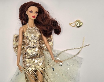May include: A fashion doll with long auburn hair and red lipstick, wearing a gold sequined halter dress with fringe. A gold heart pendant necklace and a beaded accessory are also present. The doll is posed against a white background.