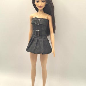 K&ouml;nnte beinhalten: Eine Puppe mit langem dunkelbraunem Haar, bekleidet mit einem tr&auml;gerlosen schwarzen Minikleid in Jeansoptik, das zwei horizontale schwarze Riemen mit silberfarbenen rechteckigen Schnallen am Oberteil sowie einen Faltenrock aufweist, dazu einfache wei&szlig;e Sneaker.