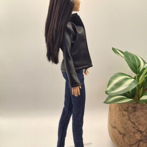 May include: A doll wearing a black faux leather jacket and dark blue denim jeans. The doll has long, dark brown hair and white shoes. The doll is standing on a clear plastic stand.