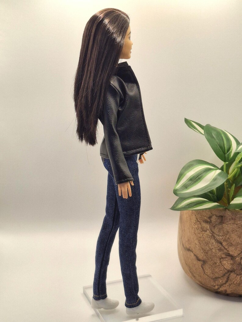 May include: A doll wearing a black faux leather jacket and dark blue denim jeans. The doll has long, dark brown hair and white shoes. The doll is standing on a clear plastic stand.
