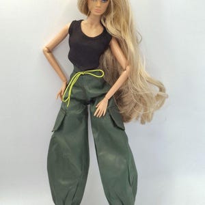 May include: A fashion doll with long blonde hair wearing a black sleeveless top and dark green cargo pants. The pants feature side pockets, a neon yellow drawstring at the waist, and elasticized cuffs at the ankles. The doll wears black boots.