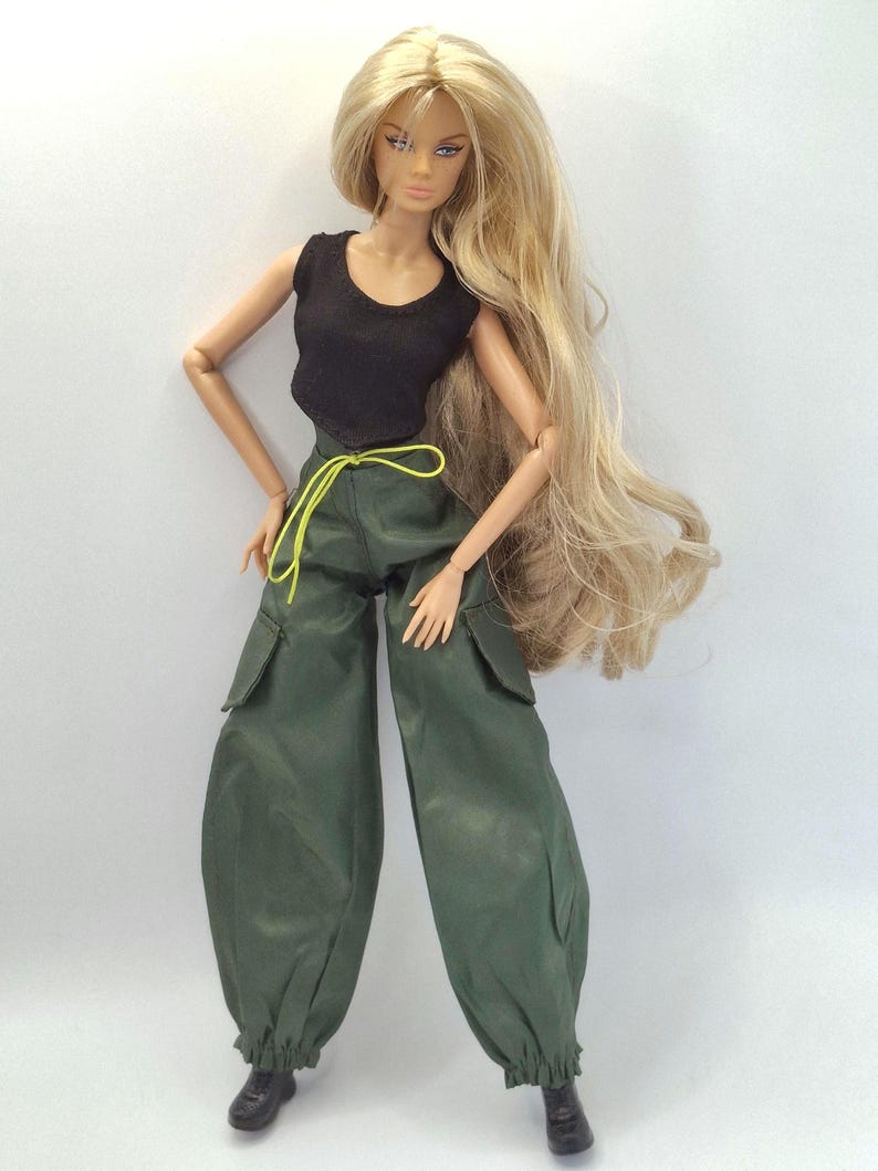 May include: A fashion doll with long blonde hair wearing a black sleeveless top and dark green cargo pants. The pants feature side pockets, a neon yellow drawstring at the waist, and elasticized cuffs at the ankles. The doll wears black boots.