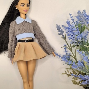 May include: A doll wearing a gray sweater with a light blue collar and a tan skirt. The doll has long black hair and is wearing black boots. A black belt with a silver buckle cinches the waist. Blue flowers are in the background.