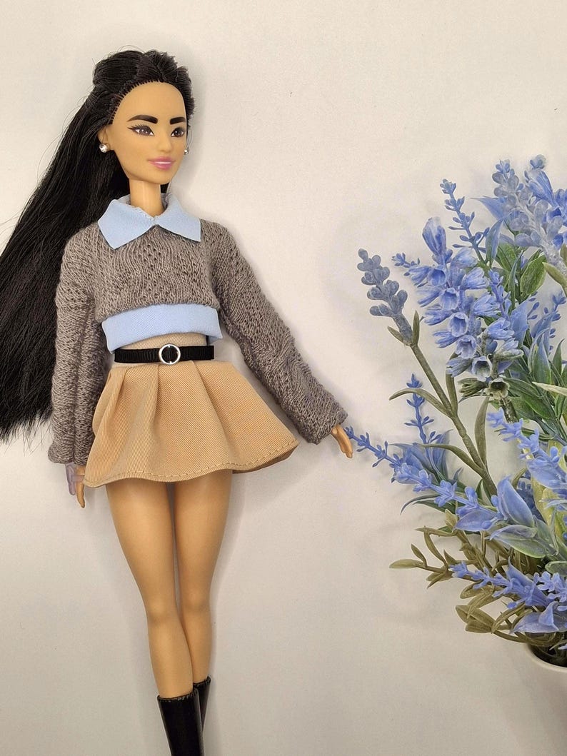 May include: A doll wearing a gray sweater with a light blue collar and a tan skirt. The doll has long black hair and is wearing black boots. A black belt with a silver buckle cinches the waist. Blue flowers are in the background.