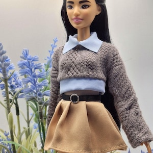 May include: A doll wearing a gray knit sweater with a light blue collar and a tan pleated skirt. The doll has long black hair and is posed in front of blue flowers.