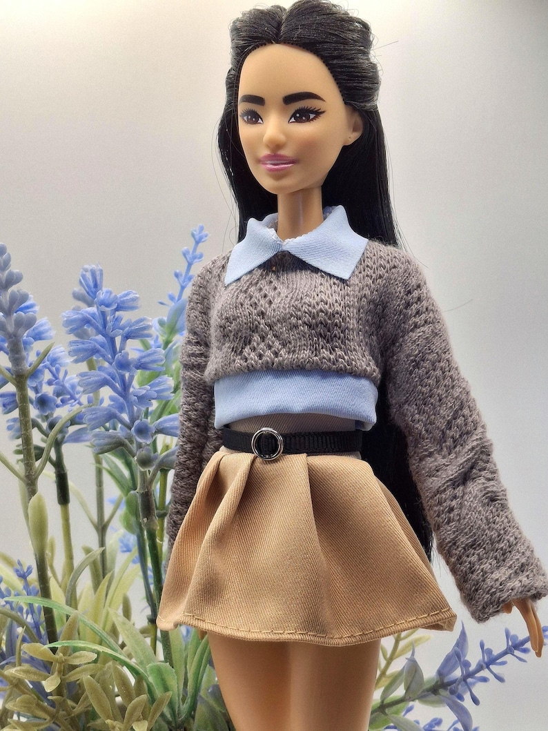 May include: A doll wearing a gray knit sweater with a light blue collar and a tan pleated skirt. The doll has long black hair and is posed in front of blue flowers.