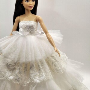 May include: A white strapless tulle wedding dress with lace detailing and a white satin bow. The dress is worn by a doll with long black hair.