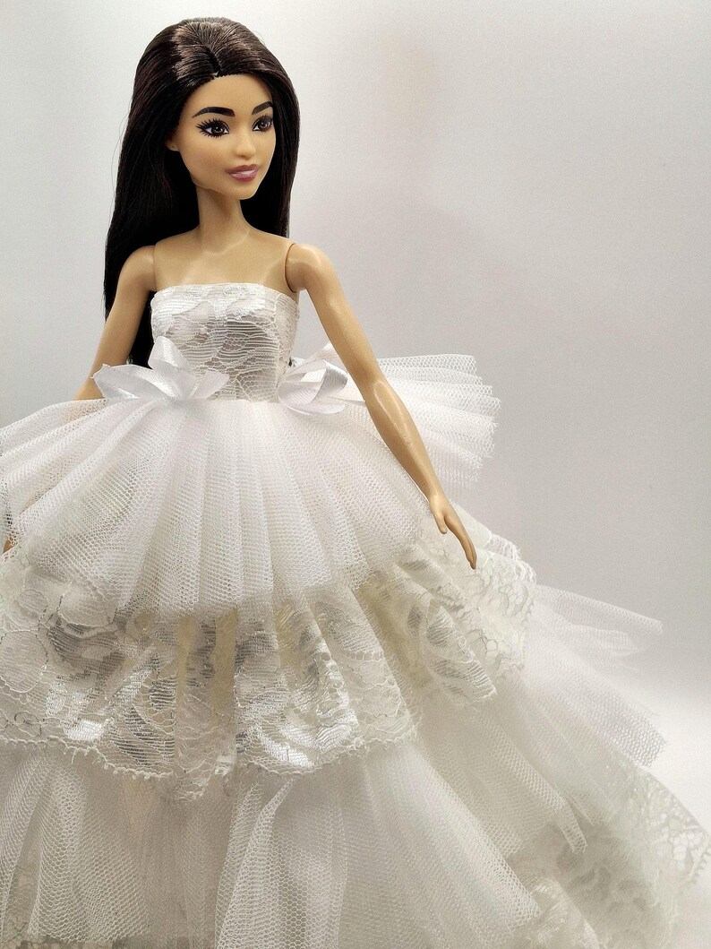 May include: A white strapless tulle wedding dress with lace detailing and a white satin bow. The dress is worn by a doll with long black hair.