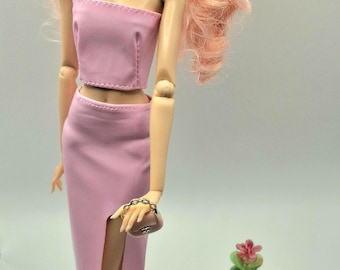 May include: A doll wearing a pink two-piece outfit, consisting of a strapless crop top and a high-slit skirt. The doll has pink hair and is holding a small brown purse with a gold chain. A small potted succulent plant is in the background.