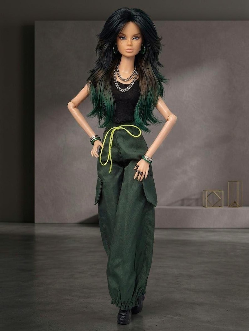 May include: A fashion doll with long dark hair featuring green highlights, wearing a black sleeveless top, dark green cargo pants with a bright yellow drawstring, silver chain necklaces, hoop earrings, and stacked bracelets on both wrists.
